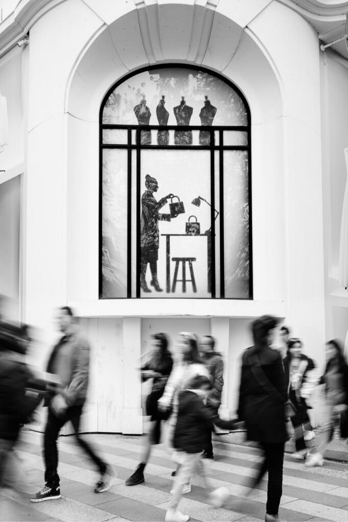 Black and white street photograph of people walking past an arched storefront window.