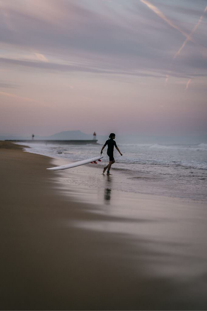 A child walking toward the ocean with a surfboard at dawn, bathed in soft early morning light.
