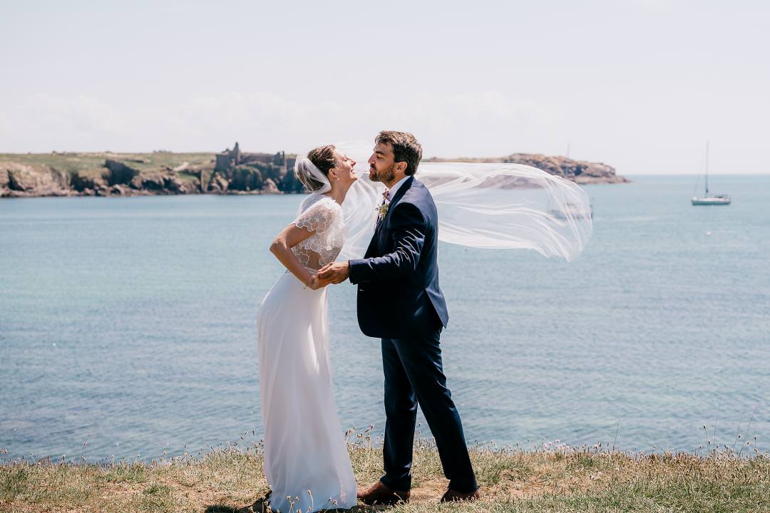 Elegant destination wedding couple standing by the sea with the bride’s veil blowing in the wind.