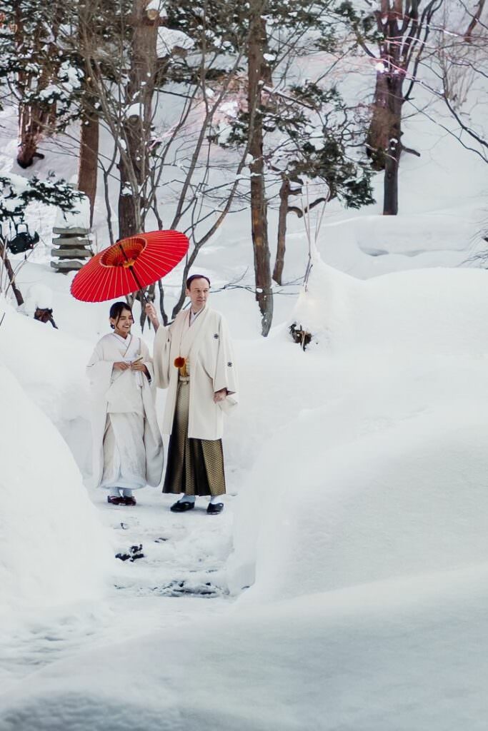 Elegant international couple standing in the snow under a red traditional umbrella during their Japanese wedding in Hokkaido.