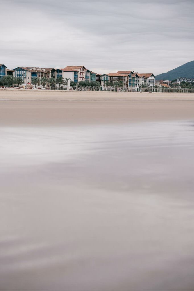 A quiet, empty beach with soft reflections and distant seaside houses beneath a gentle sky.