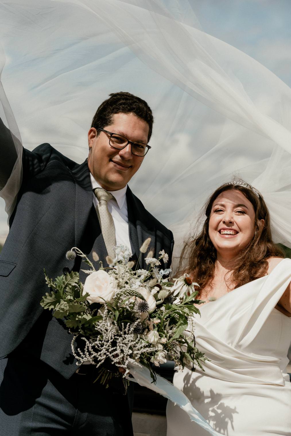 Outdoor portrait of bride and groom with the veil blowing in the wind and a floral bouquet.