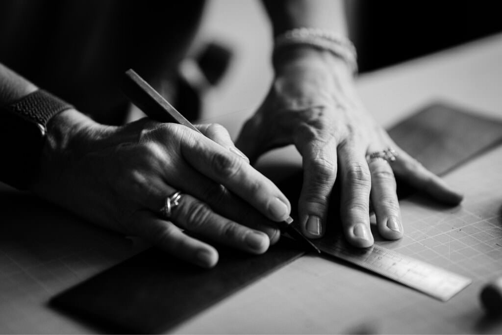 Black and white close-up of hands working with a ruler and cutter, illuminated by gentle window light.
