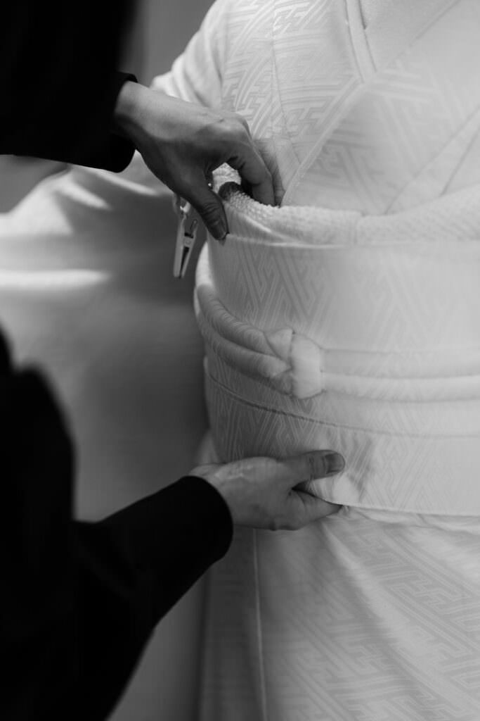 Hands tying the obi during the bride’s traditional Japanese wedding preparations.