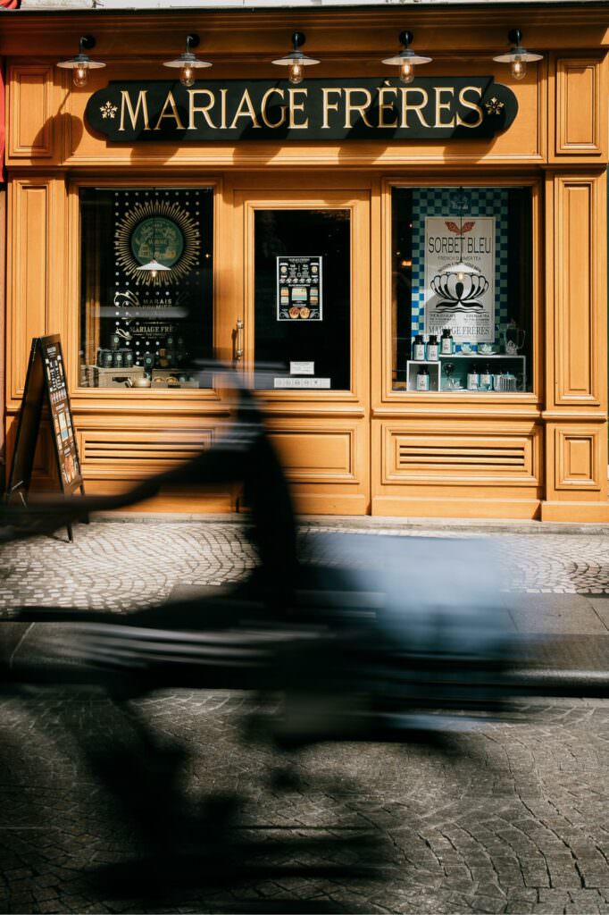 Paris street scene in front of Mariage Frères with a blurred cyclist passing in soft afternoon light.