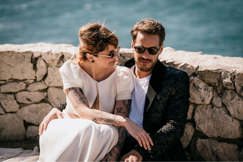 Modern wedding couple seated by the sea, captured in an editorial, fashion-inspired style.