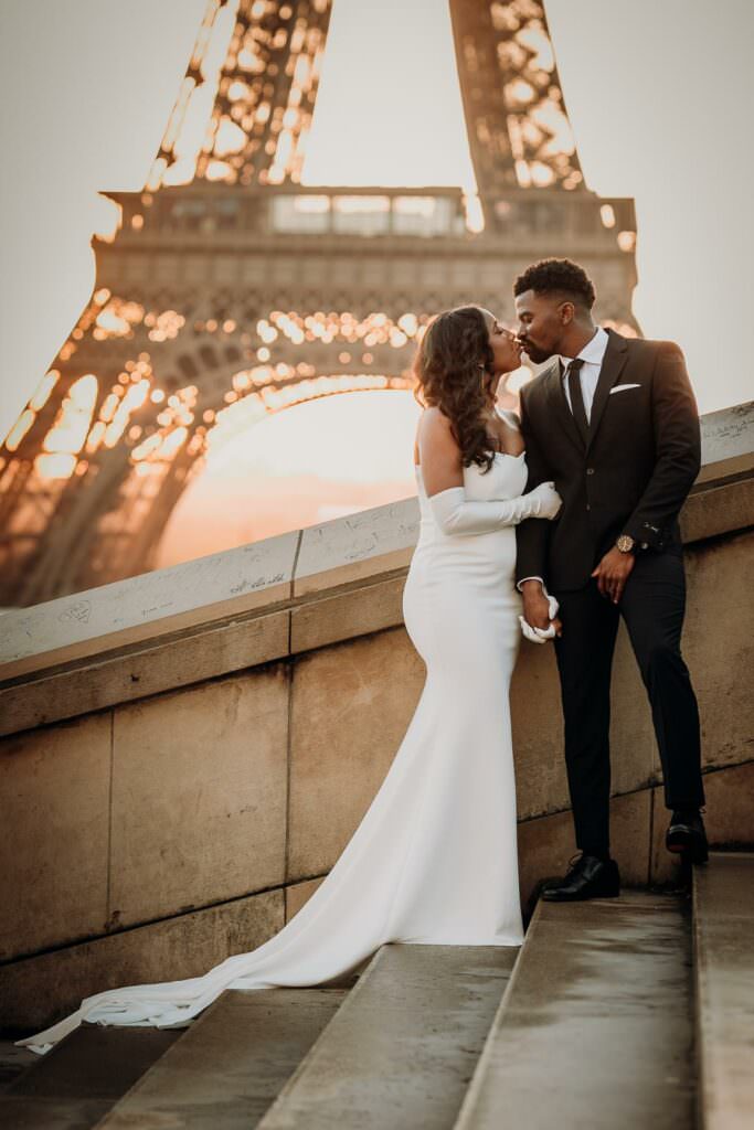 Wedding couple kissing at sunrise in front of the Eiffel Tower in Paris, dressed in elegant modern attire.