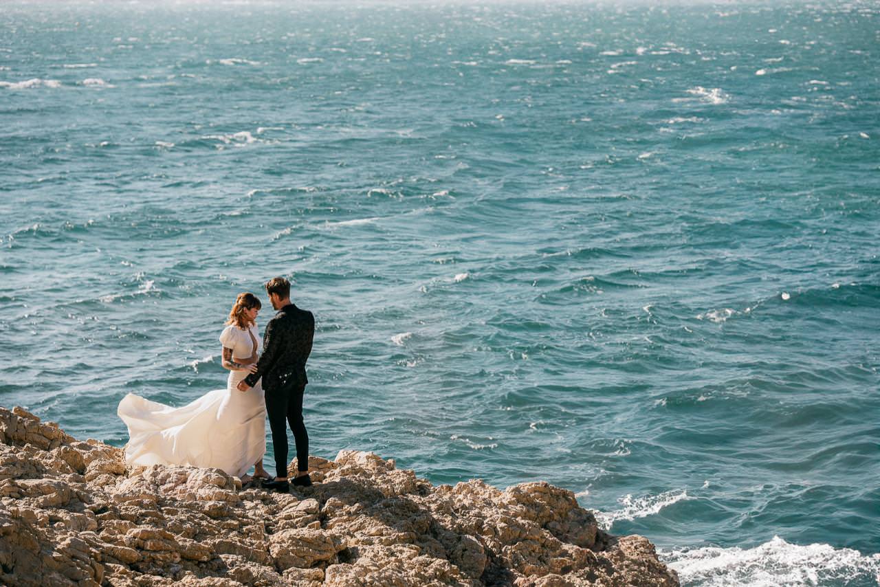 Wedding couple standing on seaside rocks in Marseille, overlooking the blue Mediterranean.